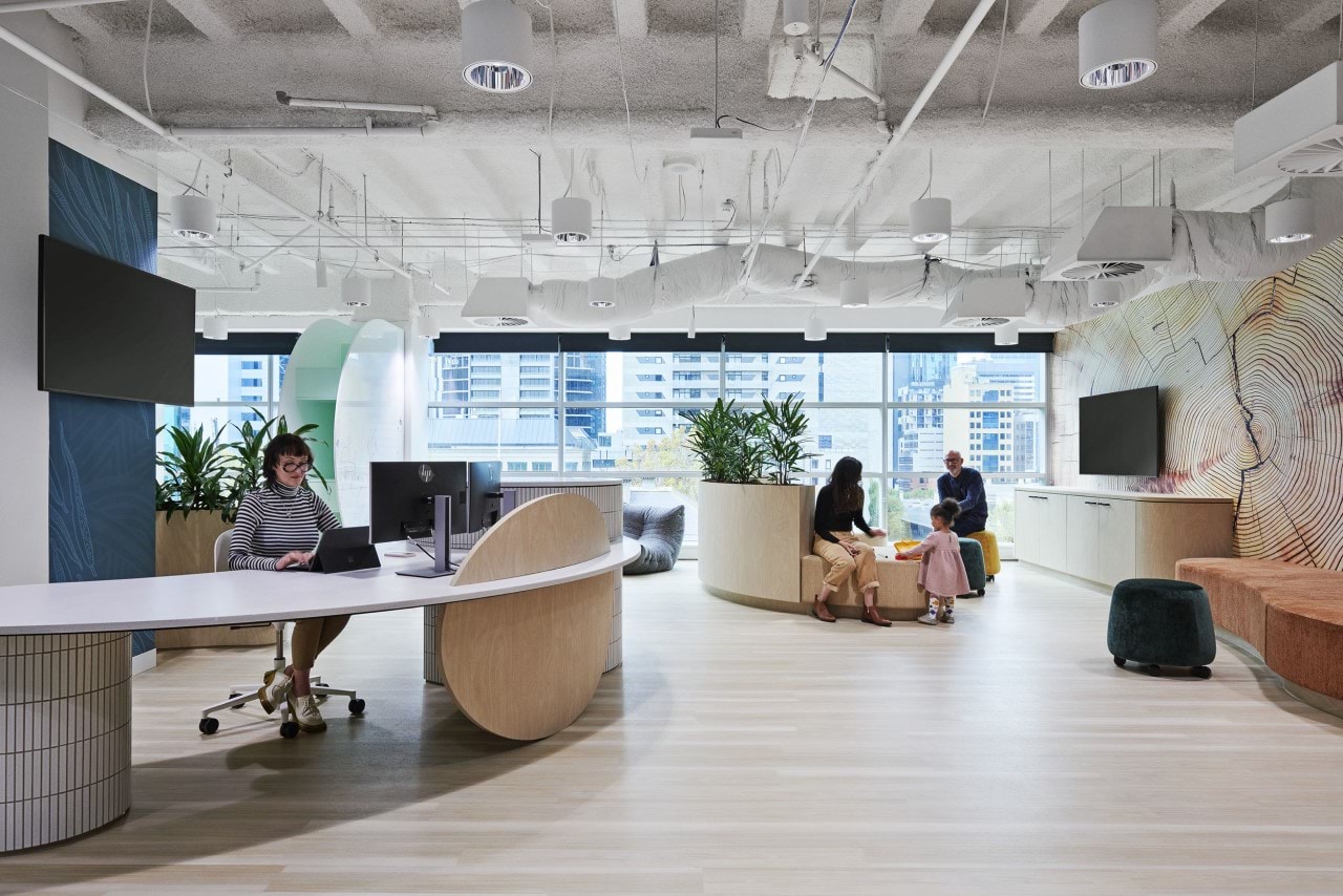 An open, modern space in a city building, with a wall of windows and exposed ceiling fittings. A receptionist sits at a moder, rounded desk, whilst a man, woman and child can be seen in the distance in the waiting area, playing with some toys. 