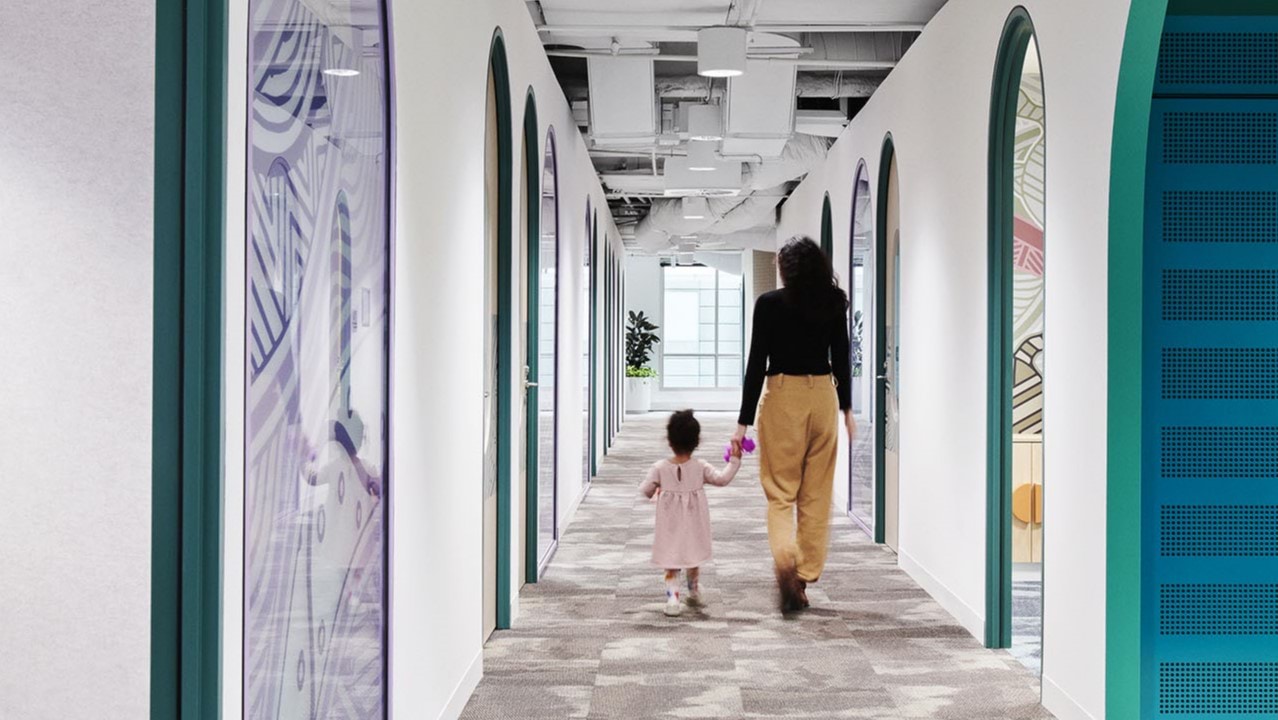 A woman holding a small girl's hand, walk away from the camera down a long, bright and airy corridor, with arched doorways on both sides, exposed ceiling and big window at the end of the corridor.